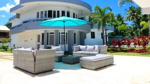 A modern outdoor seating area with wicker furniture and a blue umbrella, set against a white building with a curved roof and multiple windows. - Costa Paraíso Beach House (Aguada)
