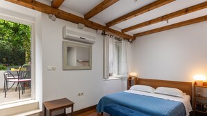 Ein Schlafzimmer mit Holzbalkendecke, einem Bett mit blauer Bettdecke, einem Nachttisch, einem Beistelltischchen mit Lampe, einem Fenster mit Vorhängen und Blick auf eine Außenpatio mit Stühlen und Tisch.