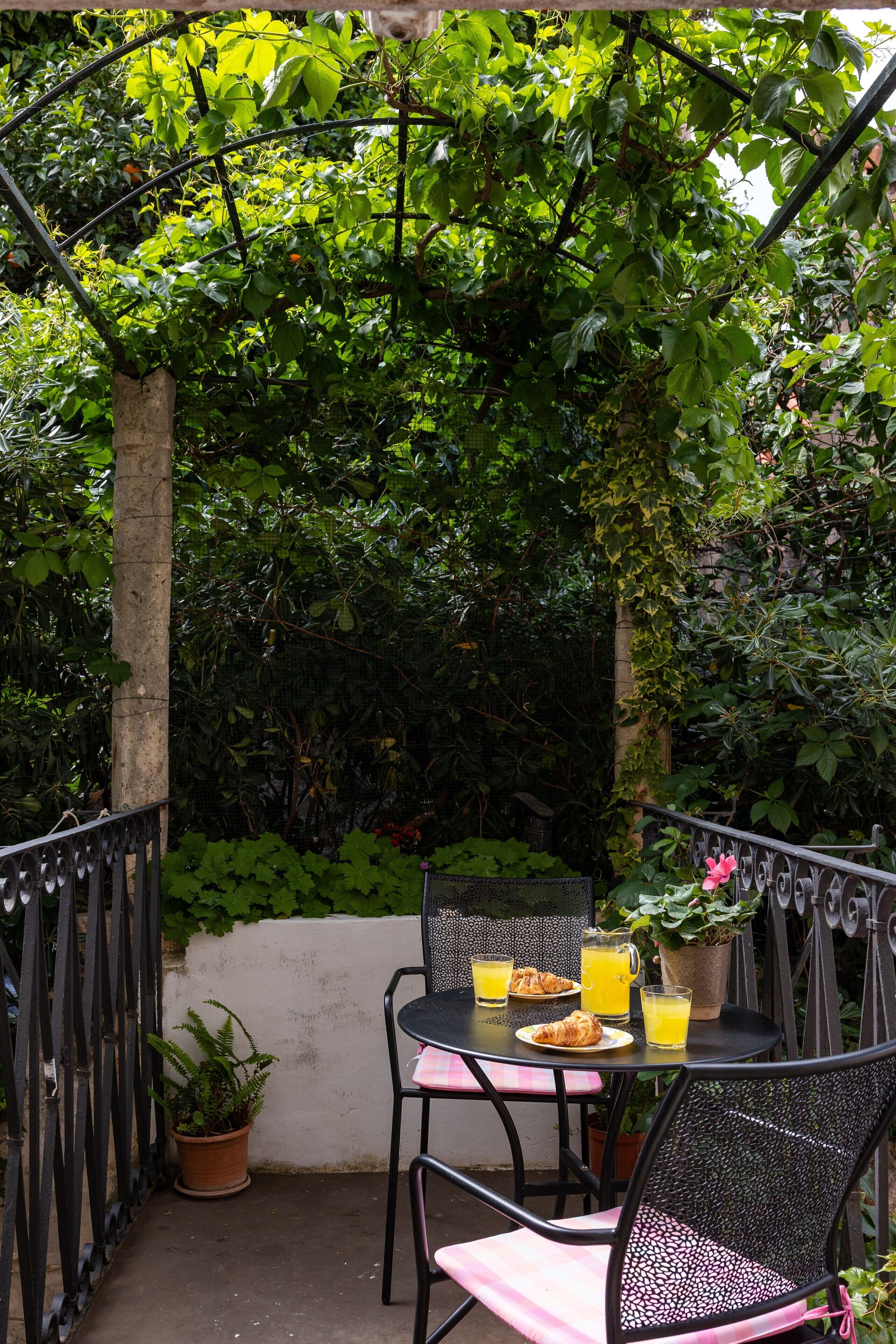 Eine Terrasse im Freien mit einem für das Frühstück gedeckten Tisch, darunter Croissants und Orangensaft, umgeben von üppiger Vegetation und einem Metallzaun.