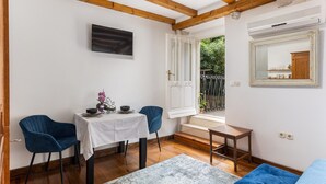 Ein Zimmer mit Holzboden, einem weißen Tisch mit weißem Tuch, zwei blauen Stühlen, einem kleinen Holztisch, einem blauen Sofa, einem Spiegel und einem Fenster mit Blick auf Grünflächen.