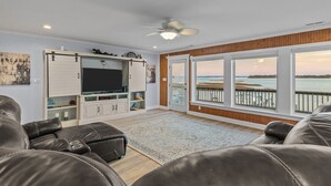 Ein geräumiges Wohnzimmer mit großem Fernseher, Ledersofas und Blick auf das Wasser durch große Fenster.
