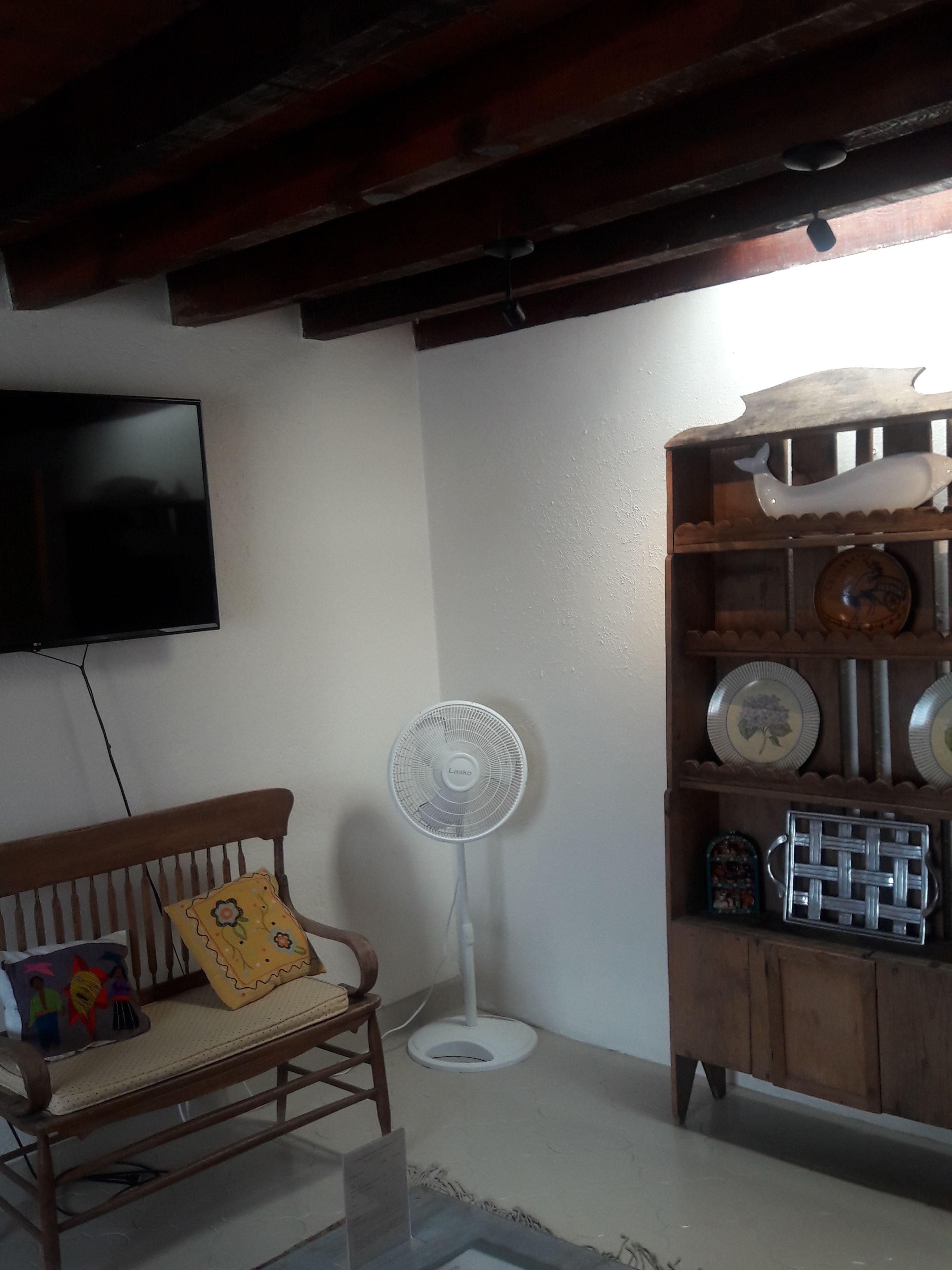 A room with a wooden bench, a standing fan, a television mounted on the wall, and a wooden cabinet with decorative plates.