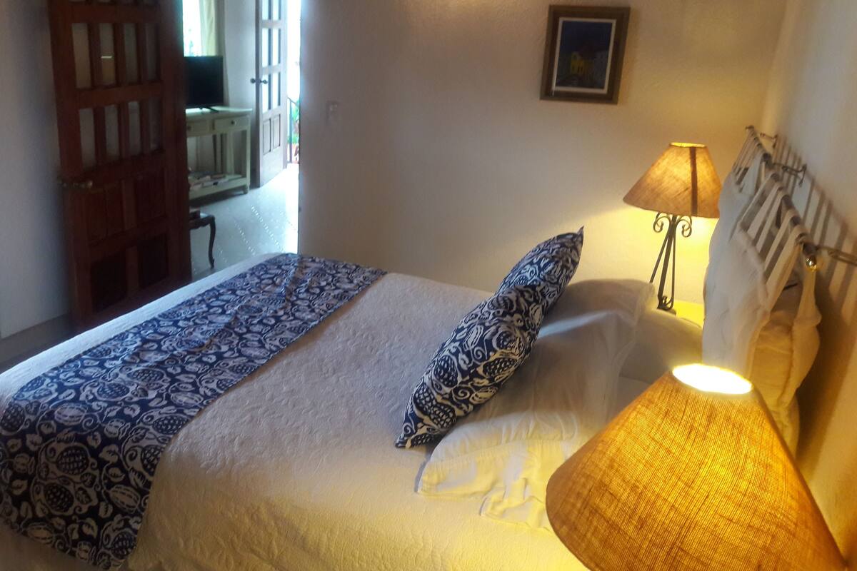 Une chambre à coucher avec un lit, une literie à motifs, une lampe de chevet, un tableau encadré au mur et une porte donnant sur une autre pièce.