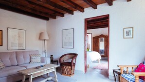 Una sala de estar acogedora con un sofá, una mesa de centro de madera, un sillón de mimbre y un banco de madera. Hay cuadros enmarcados en las paredes y una puerta de madera que da a otra habitación.