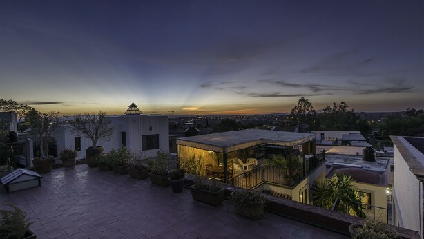 Una terraza en la azotea con plantas en macetas, un espacio de descanso y vistas al horizonte de la ciudad al atardecer.