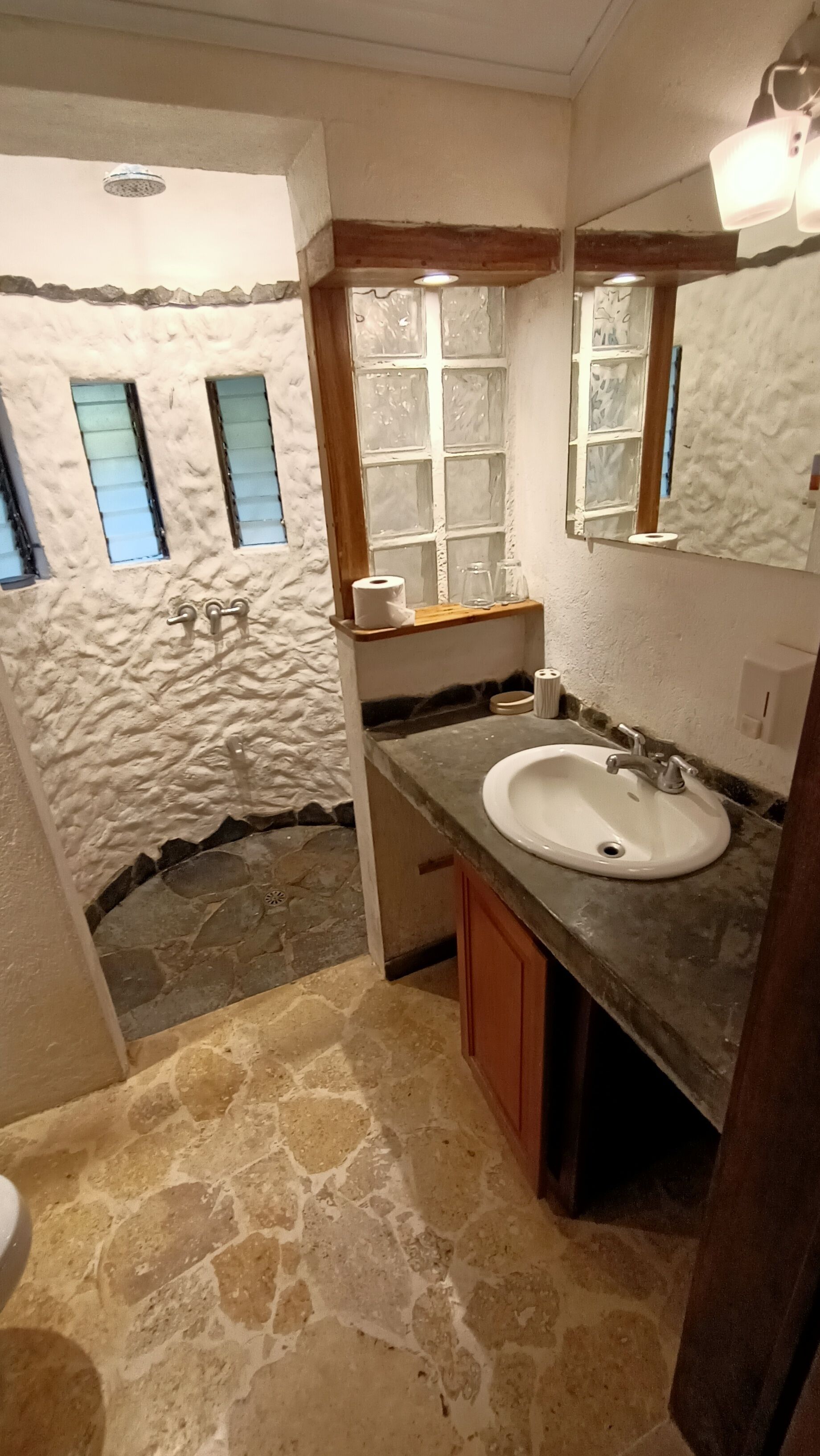 Un baño con suelo de piedra, un lavabo blanco, armarios de madera, un espejo y una ventana con vidrio esmerilado.