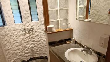 Un baño con suelo de piedra, un lavabo blanco, armarios de madera, un espejo y una ventana con vidrio esmerilado.