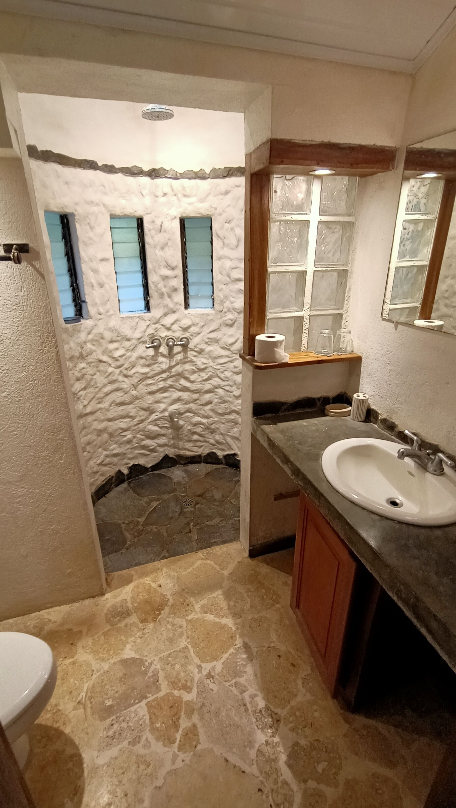 Une salle de bain avec un sol en pierre, un lavabo blanc et une fenêtre avec des volets bleus.
