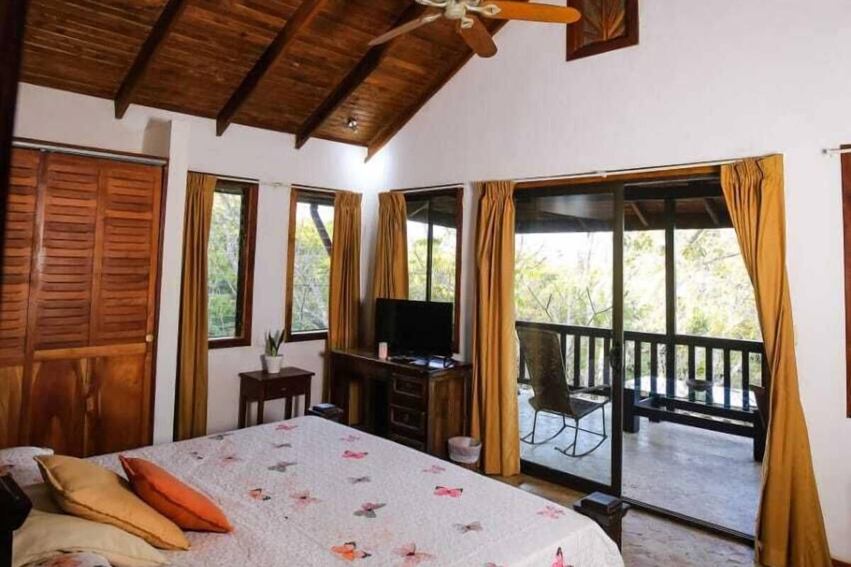 Un dormitorio con techo de madera, cama con colcha floral, escritorio con televisor y balcón con cortinas.