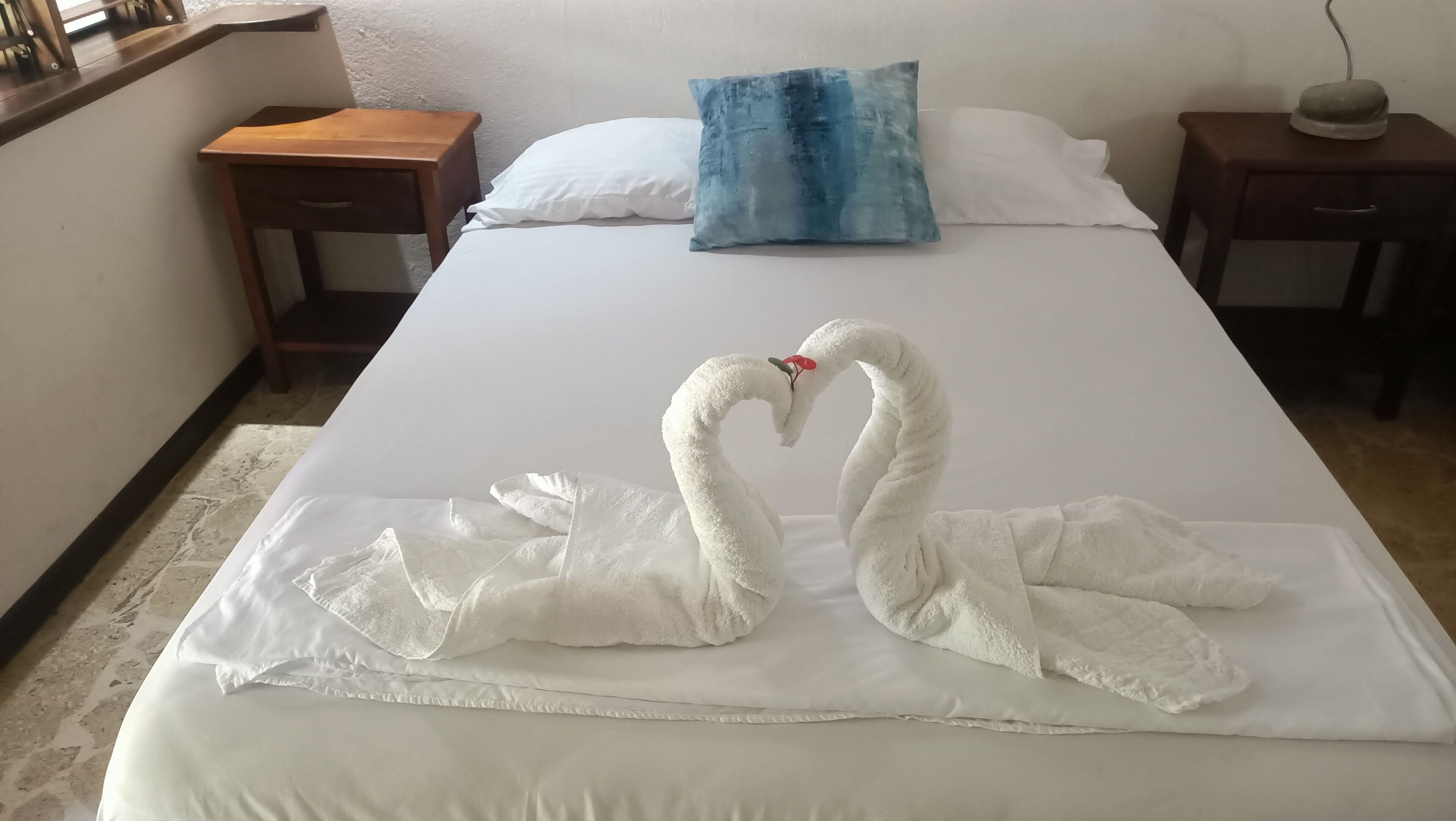 Una cama impecable con sábana y dos toallas blancas dispuestas en forma de corazón.