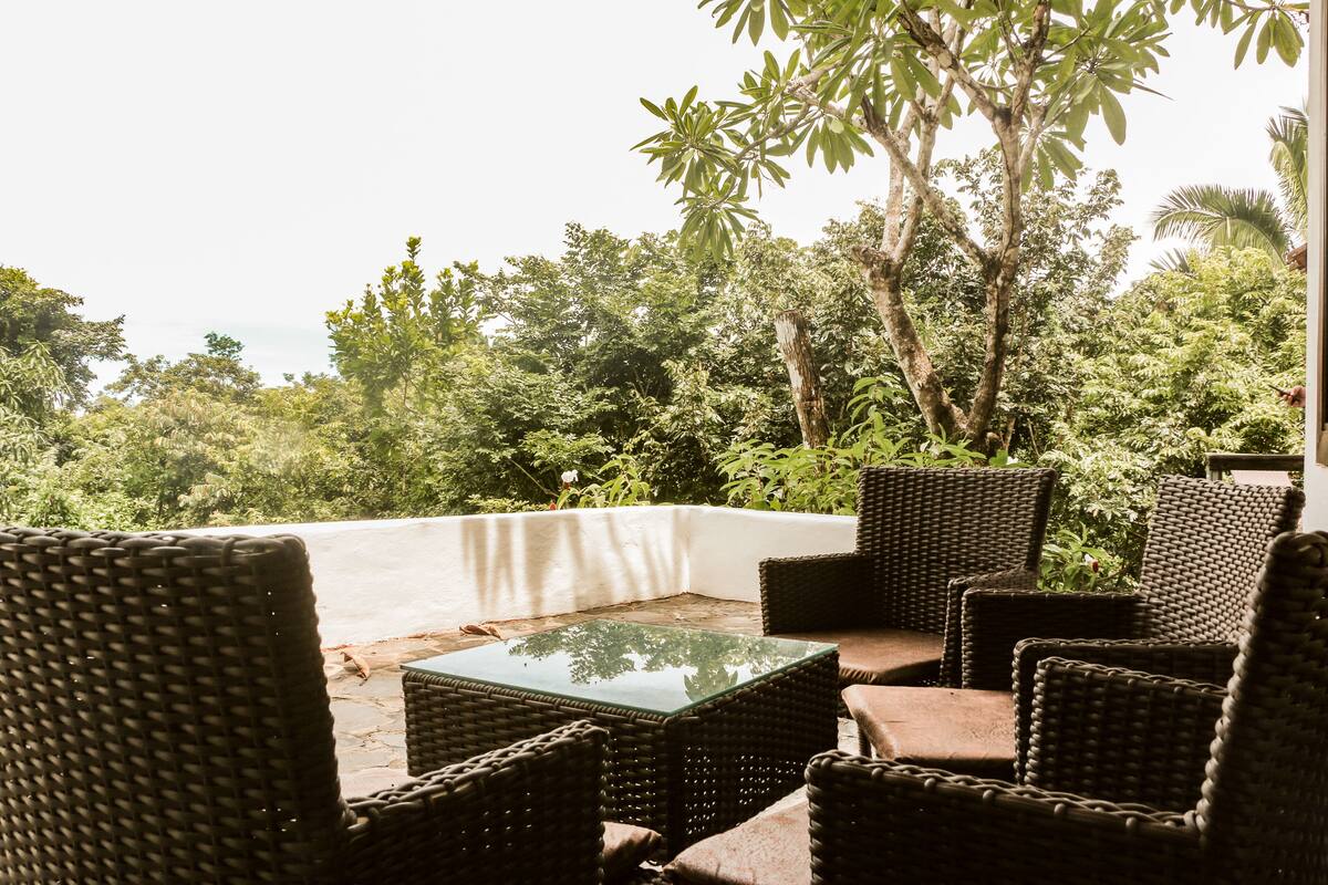 Un área de estar al aire libre con mobiliario de mimbre y una mesa de vidrio, con vistas a un exuberante jardín y el océano.