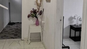 Un pasillo con una puerta blanca, una pequeña mesa blanca con un jarrón de flores, un cuadro en la pared y una mesa negra con una lámpara.