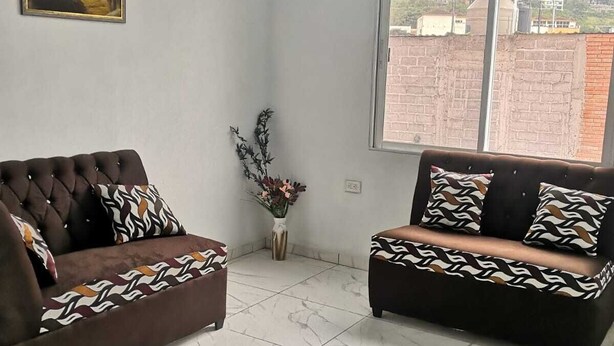 Una habitación con dos sofás a juego y un reposapiés, un cuadro en la pared y un ventanal con vistas a edificios.