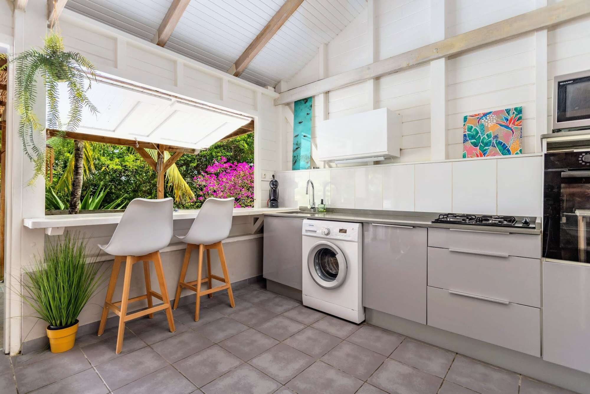 A compact kitchen with a washing machine, a stove, and a sink. There are two bar stools and a window overlooking a garden with pink flowers.