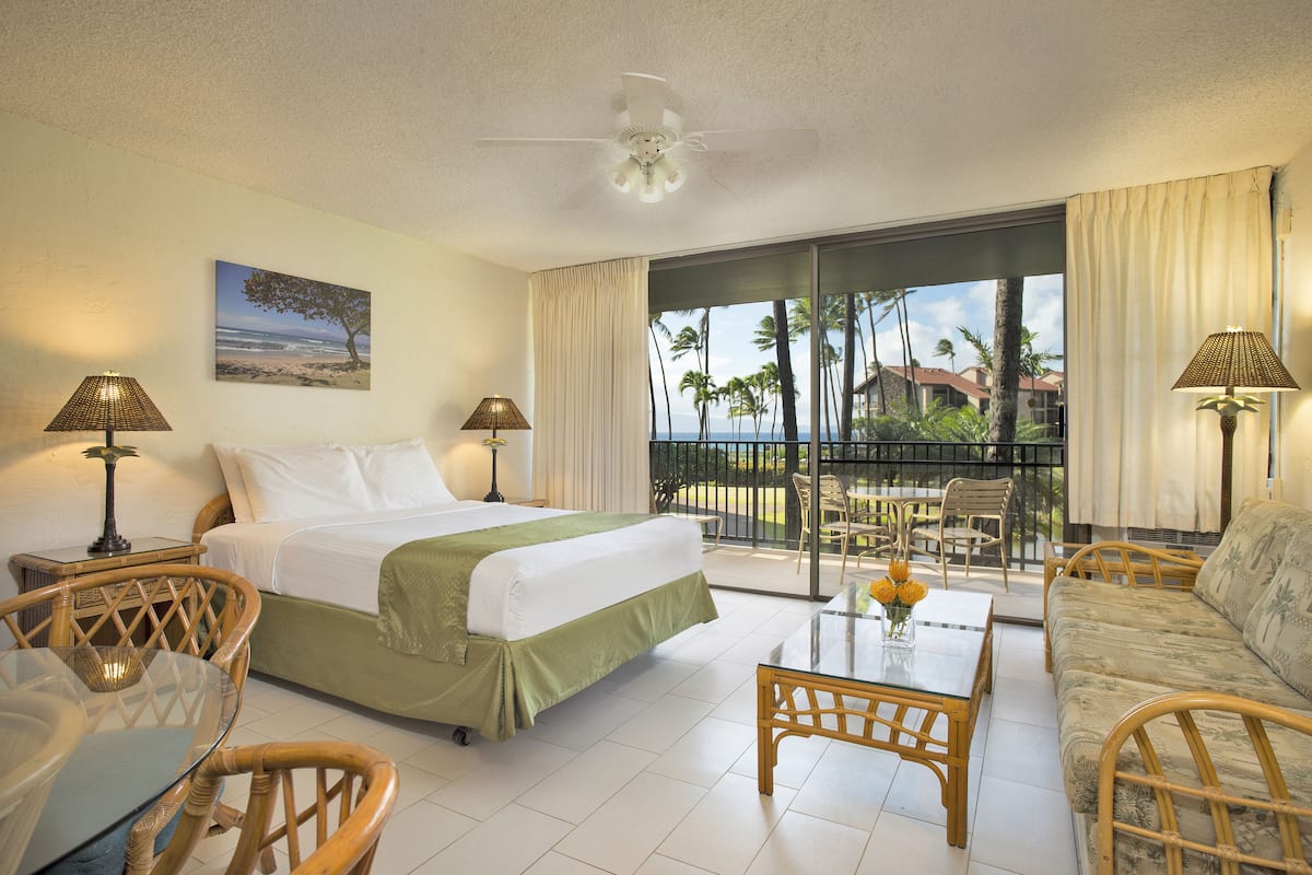 A hotel room with a bed, two chairs, a table, a balcony with a view of palm trees and a beach.