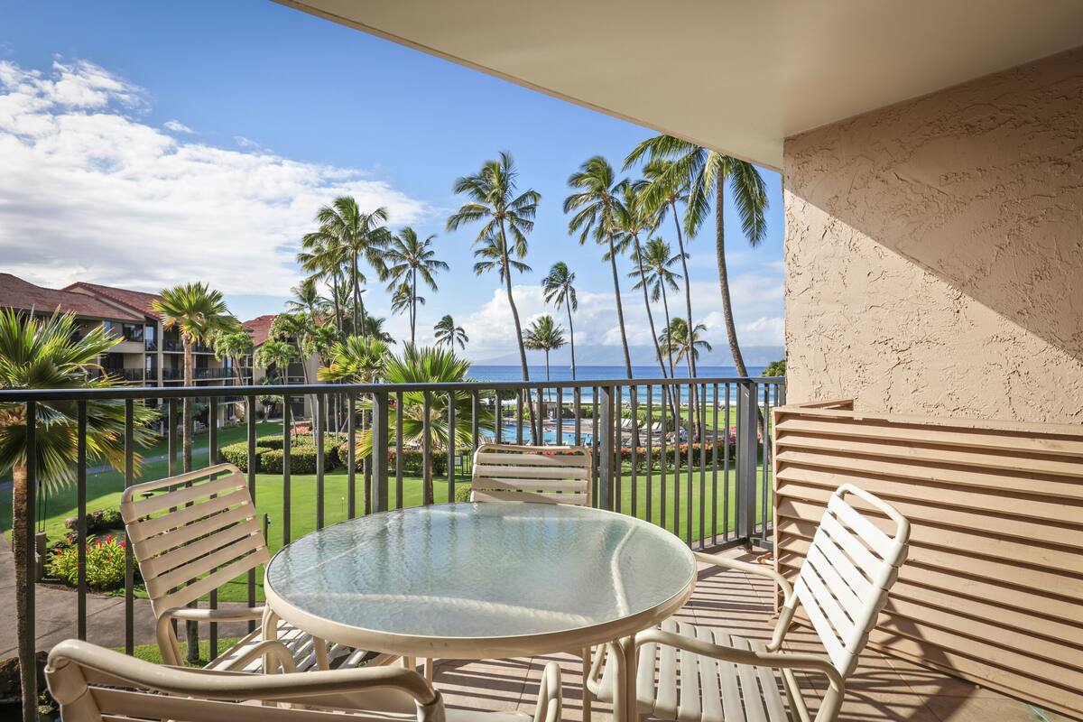 A balcony with a glass table and chairs, overlooking a pool and palm trees with a view of the ocean.