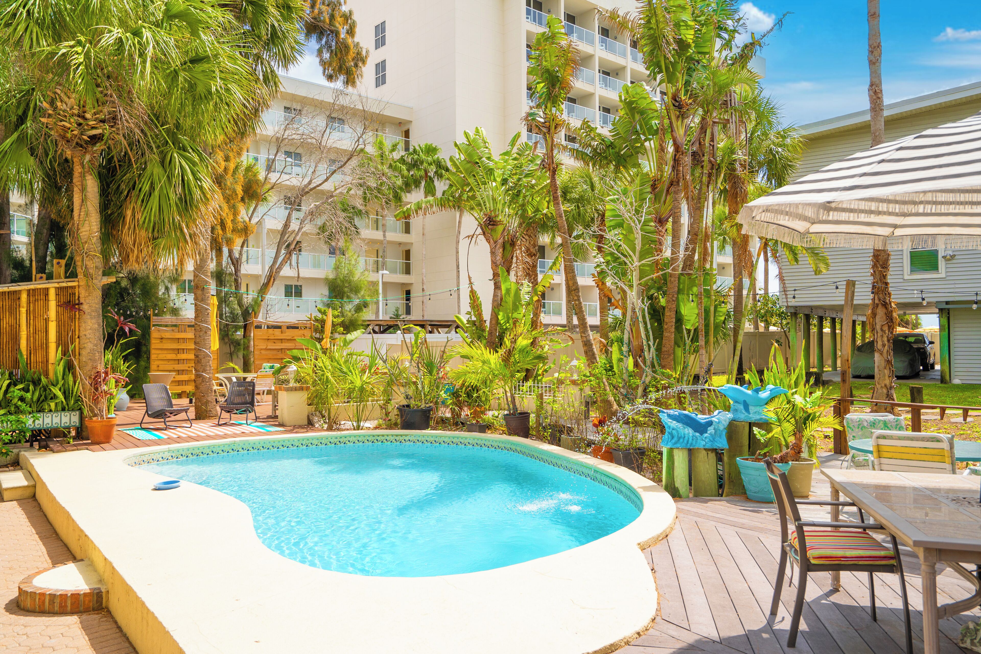 A swimming pool surrounded by palm trees and potted plants, with outdoor furniture and a building in the background.