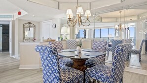 Een eetruimte met een ronde tafel, omringd door stoelen met een patroon, een kroonluchter en een grote spiegel die de kamer weerspiegelt.