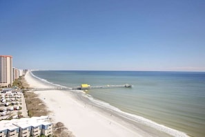 A beach with a pier extending into the water, adjacent to a row of buildings.