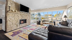 Un salon moderne avec une cheminée en pierre, une grande fenêtre donnant sur les arbres aux couleurs automnales et un canapé en cuir.