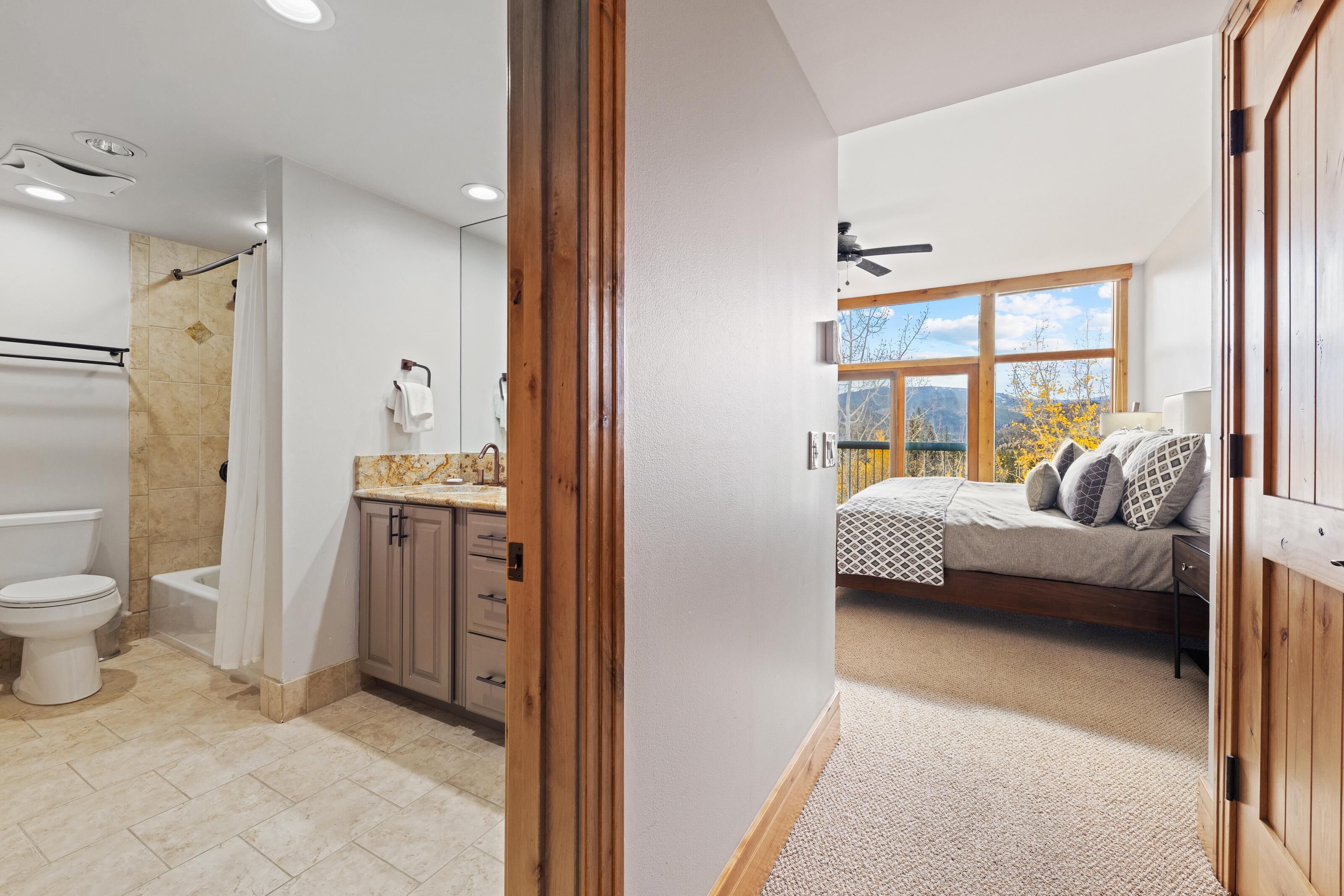 Ein modernes Badezimmer mit Dusche, Toilette und Waschtisch. Durch eine geöffnete Tür ist ein Schlafzimmer mit Bett, Fenster und Blick ins Freie zu sehen.