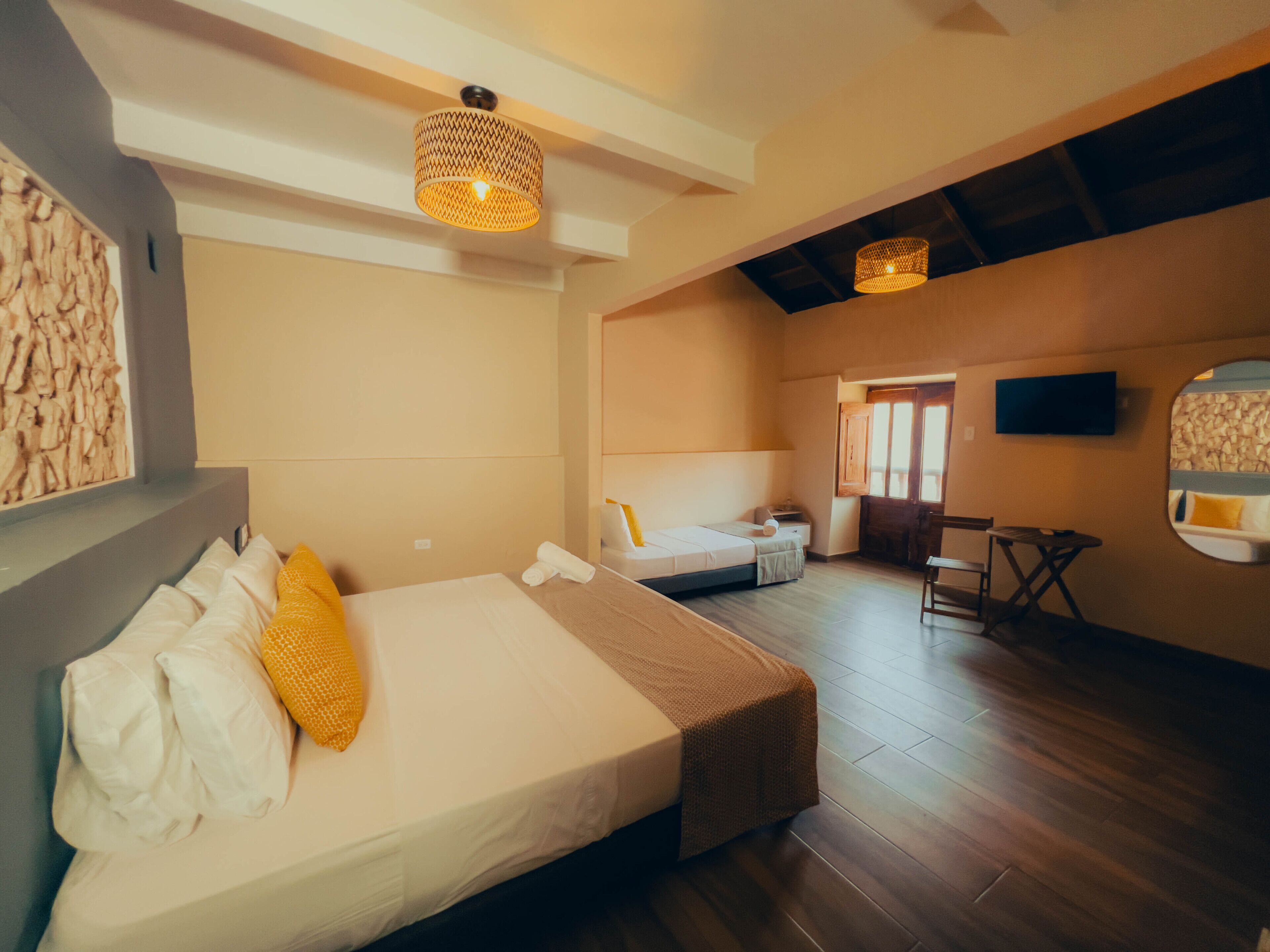 A hotel room with a large bed, a smaller bed, a desk, and a chair. There is a television mounted on the wall and a window with a view of the outside.