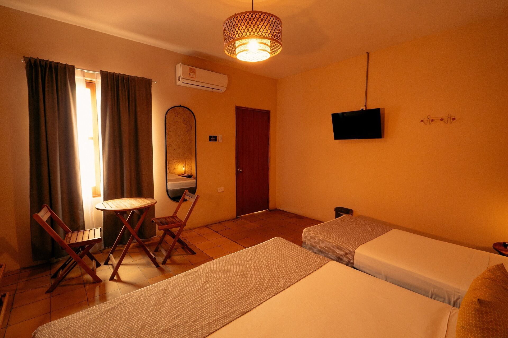 A hotel room with two beds, a small table, a chair, a mirror, an air conditioning unit, and a television.