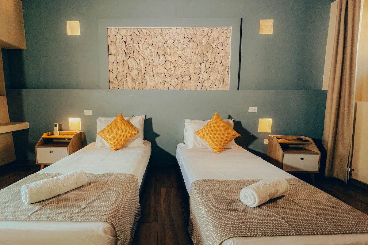Een hotelkamer met twee bedden, elk voorzien van een witte deken en een bruine gebreide plaid. Twee nachtkastjes met lampen, een wanddecoratie en een houten kastje zijn aanwezig.