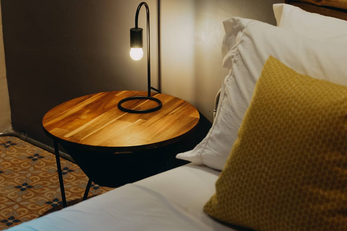 Een bed met witte beddengoed, een rond houten nachtkastje met een lamp, en een vloer met een patroon.