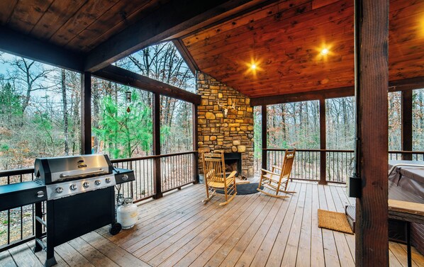 A cabin with a stone fireplace, a grill, and a deck with chairs.