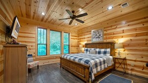 A wooden cabin bedroom with a bed, a ceiling fan, a nightstand, a lamp, a bench, and a window with blinds.