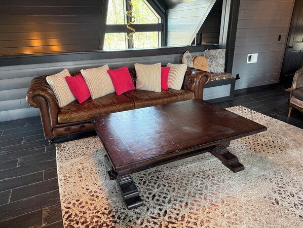 A brown leather sofa with red and beige cushions, a wooden coffee table with wheels, and a patterned rug on a dark wooden floor.