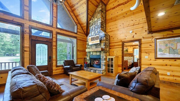 A cozy log cabin interior with a stone fireplace, leather sofas, and large windows offering a view of the outdoors.
