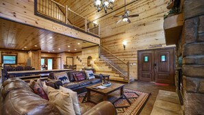 A spacious log cabin interior with a leather sofa, a dining area, a staircase, and a stone fireplace. - Caddy Shack | Luxury Group Lodge Near Broken Bow Lake with Game Room and Expansi (Broken Bow)