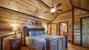 A wooden cabin bedroom with a bed, nightstands, a ceiling fan, and a wooden door. - Caddy Shack | Luxury Group Lodge Near Broken Bow Lake with Game Room and Expansi (Broken Bow)