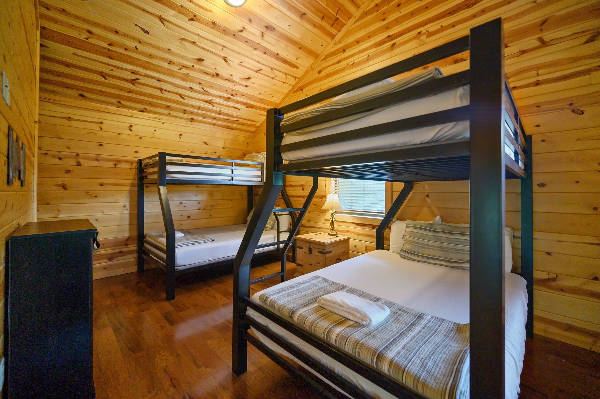 A cabin with two bunk beds, wooden walls, a small wooden table, and a window with blinds.