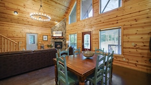 A wooden cabin interior with a dining table, chairs, a stone fireplace, and a sign reading "BROKEN BOW Cabin Lodging."