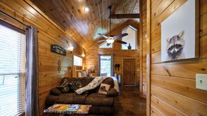 Interior acogedor de una cabaña de troncos, con techo de madera, ventilador de techo, sofá con una manta y un cartel de un mapache en la pared.