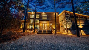 A modern two-story wooden house with a stone facade, surrounded by trees and a gravel driveway.