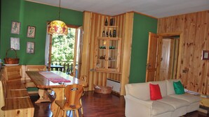 Een houten eettafel met stoelen, een bank met kleurrijke kussens en een houten kast met flessen.