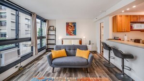 Eine moderne Wohnung mit grauem Sofa, gelben Kissen, einem bunten Teppich, einem Gemälde, einer Küche mit Holzschränken und einem großen Fenster mit Blick auf Gebäude.