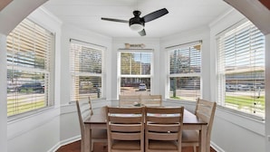 Comedor con una mesa y sillas, ventilador de techo y ventanas grandes con persianas.