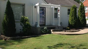 A single-story house with a front porch, a garage door, and a small front yard with two evergreen trees.