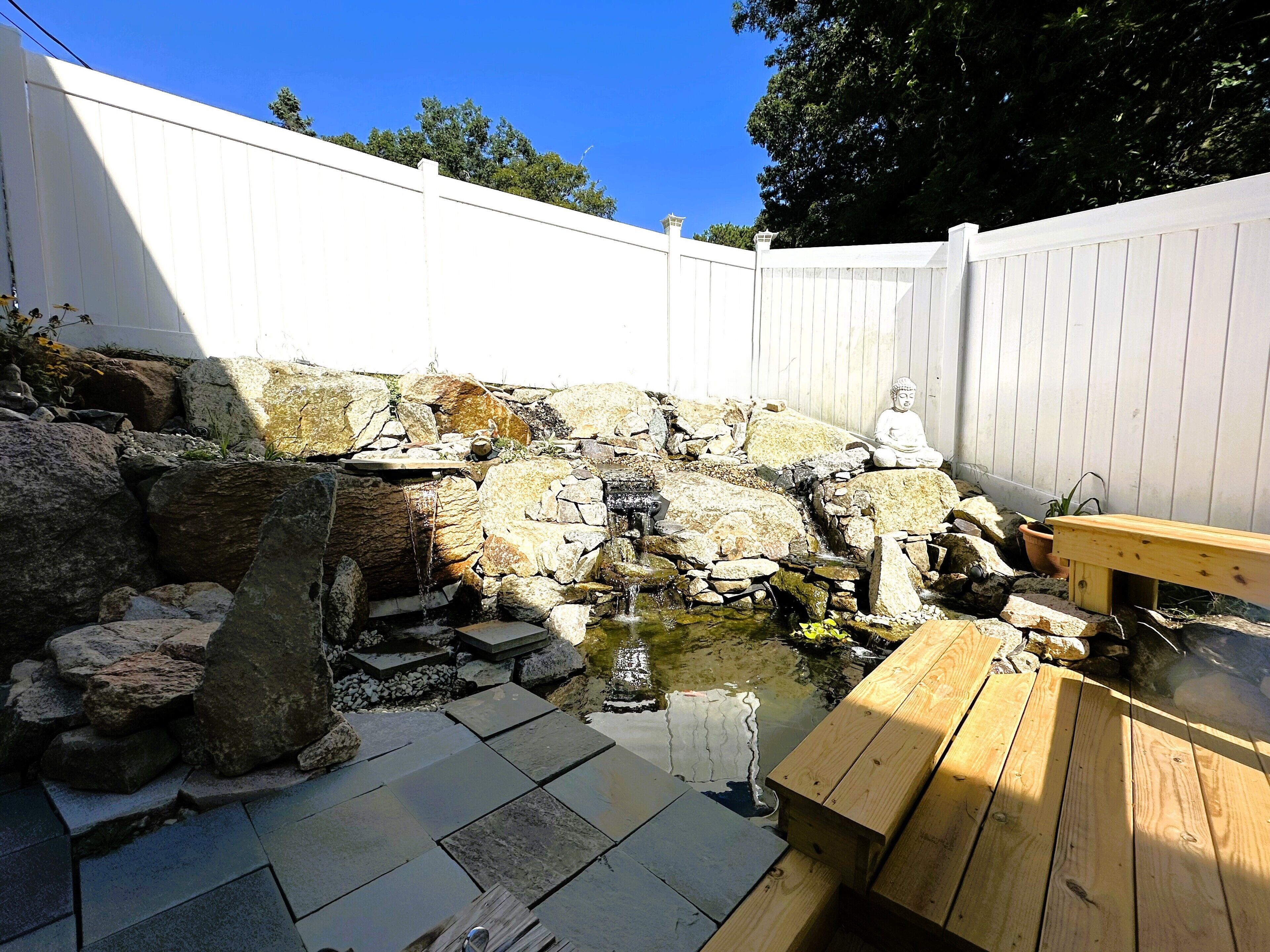A backyard with a small pond, rocks, a wooden bench, and a Buddha statue.