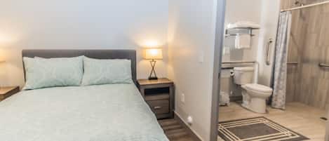 A bedroom with a bed, nightstand, lamp, and a view into a bathroom with a shower and toilet.