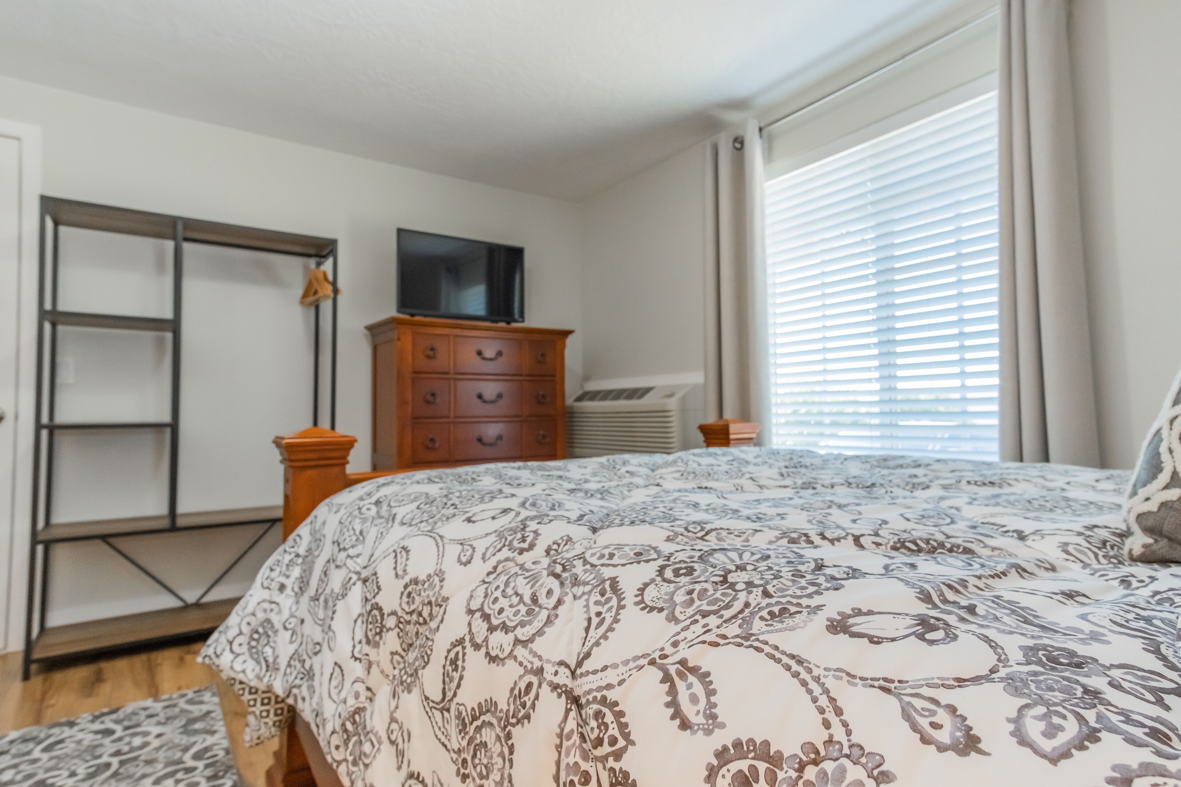 A bedroom with a bed, wooden dresser, TV, and a window with blinds.