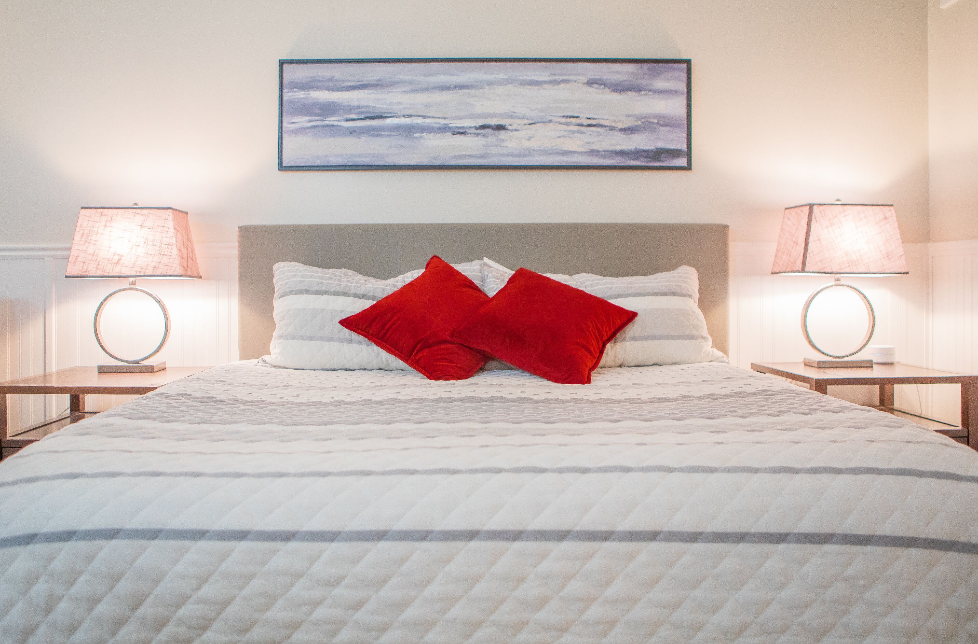 A neatly made bed with a white quilt, two red pillows, and a headboard. There are two bedside tables with lamps and a framed artwork above the bed.