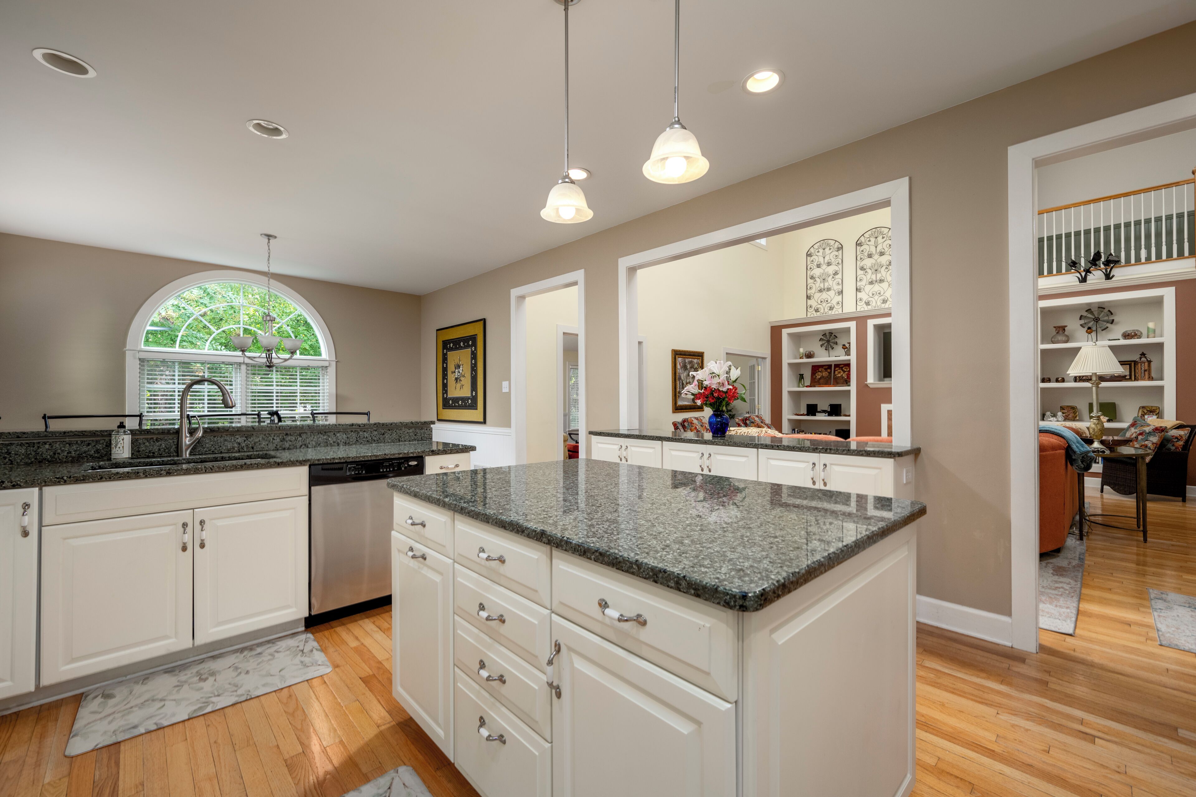 A spacious kitchen with granite countertops, white cabinetry, and a large island. There are pendant lights above the island, and a window with blinds on the left side.