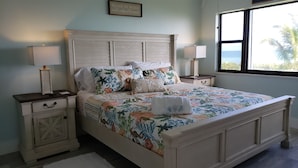 A bedroom with a bed, two bedside tables, a nightstand, a window with a view, and a framed quote on the wall. - Escape to your HAPPY place, luxury corner condo right on the beach! (Jensen Beach)