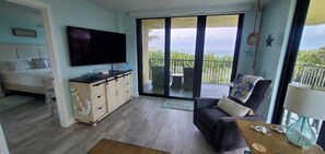 A room with a bed, a TV, a desk, and a balcony with a view of the sea.